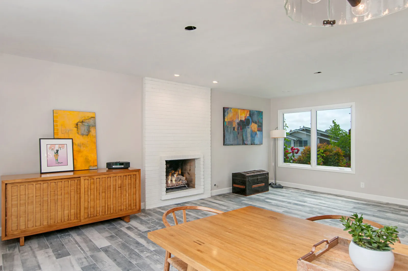A staged living room with a white painted brick fireplace, mid-century credenza, and gray wood-look flooring.